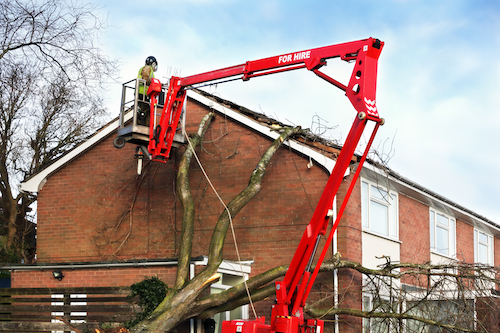 Tree surgeon working up cherry picker repairing storm damaged roof after an uprooted tree fell on top of a residential house