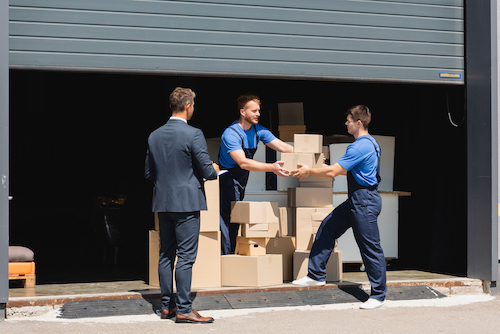 Businessman with clipboard standing near two male movers holding cardboard boxes just outside warehouse